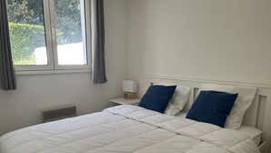 A neatly made bed with white bedding and blue pillows, a bedside table with a lamp, and a window with curtains. - Vaux-sur-Mer: 2-Bed Villa for 5, 450m to Beach, Parking, AirCon (Vaux-sur-Mer)