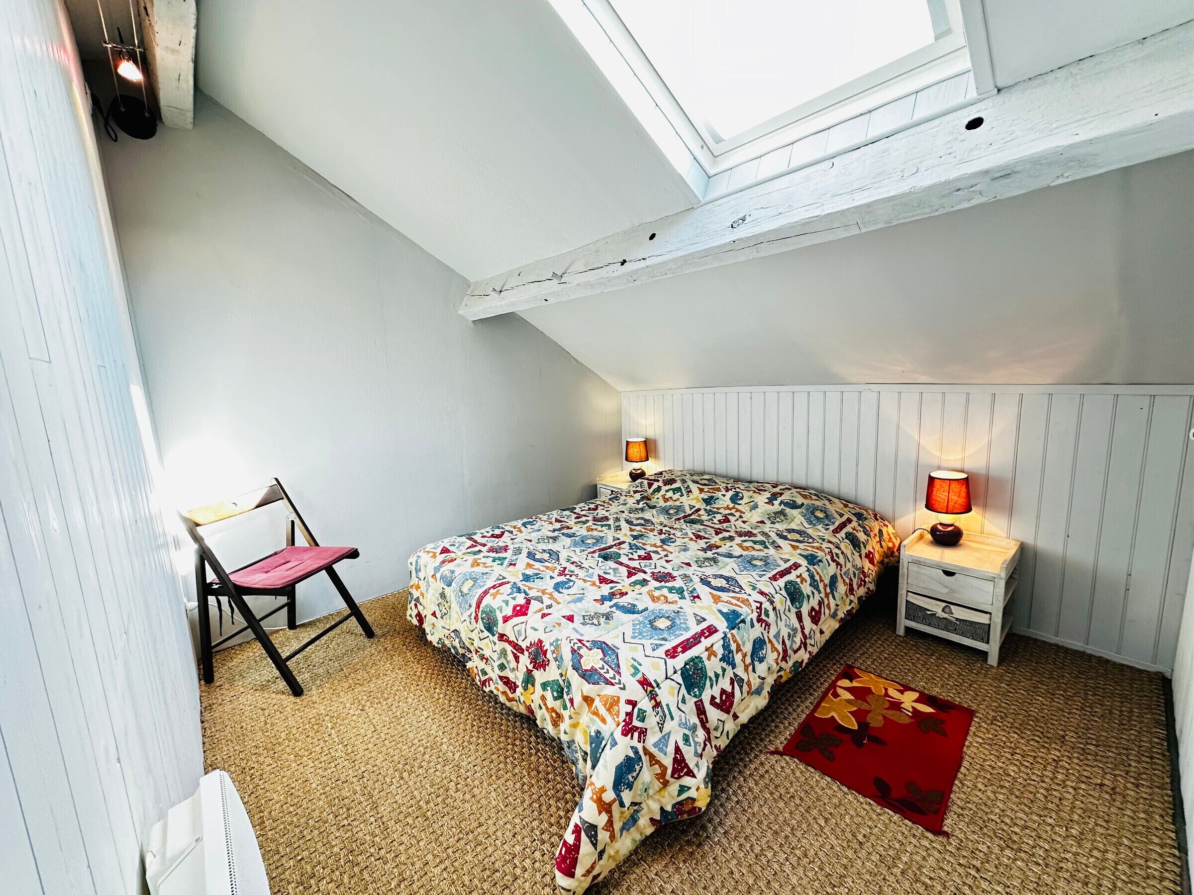 Un dormitorio pequeño y acogedor con techo inclinado, una cama con colcha de colores, dos lámparas de noche, una silla y una alfombra pequeña.