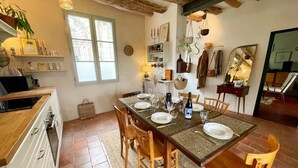 Un comedor acogedor con una mesa de madera preparada para cuatro, rodeada de sillas de madera. Se ve una cocina con estufa y armarios a la izquierda.