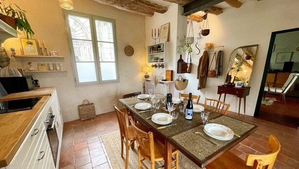 A cozy dining area with a wooden table set for four, surrounded by wooden chairs. A kitchen with a stove and cabinets is visible to the left.