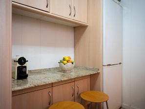 A kitchen with wooden cabinets, a granite countertop, a fruit bowl, a coffee maker, and two stools. - Apartment 6 people, pool access, private parking, near beach (Roda de Berà)