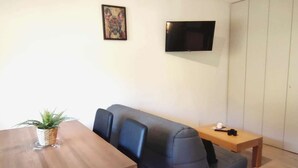 Un salon moderne avec une table en bois, un canapé bleu, une petite table d’appoint, un téléviseur fixé au mur et une plante en pot.