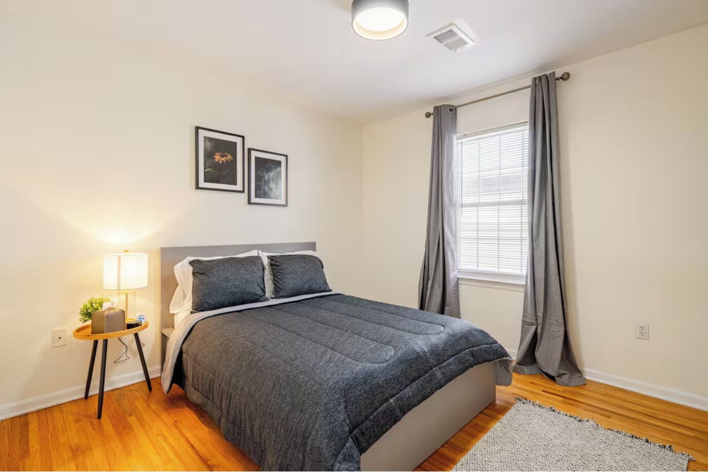 Un dormitorio con cama, mesita de noche con lámpara, dos cuadros enmarcados en la pared, una ventana con cortinas y suelo de madera.