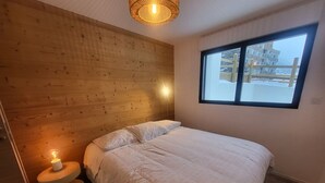 A bedroom with a wooden headboard, a bed with white bedding, a bedside lamp, and a window with a view of buildings. - Montgenèvre Apartment with Terrace, Wifi & Parking (Montgenèvre)