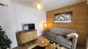 A living room with a television on a wooden cabinet, a grey sofa, a wooden coffee table with glasses and a bottle, a Christmas tree, and a wall-mounted picture of polar bears. - Montgenèvre Apartment with Terrace, Wifi & Parking (Montgenèvre)