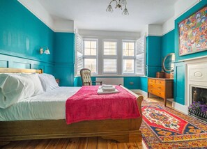 A bedroom with a bed, a chair, a dresser, a fireplace, and a window with blinds.