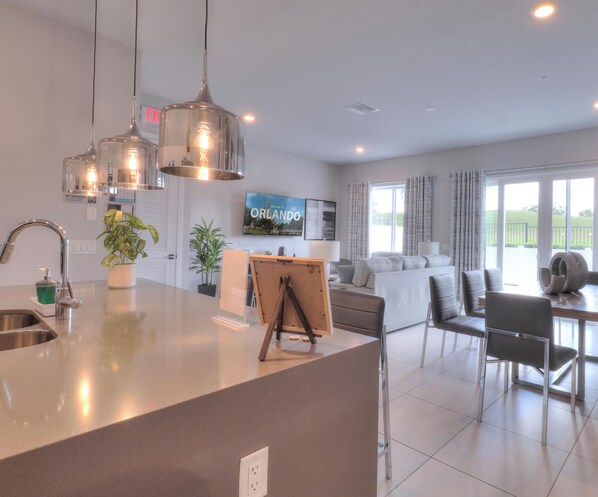 A modern kitchen with a central island, dining area with chairs, and a living room with a sofa and TV. - Modern Condo in Reunion Resort Near Disney (Kissimmee)