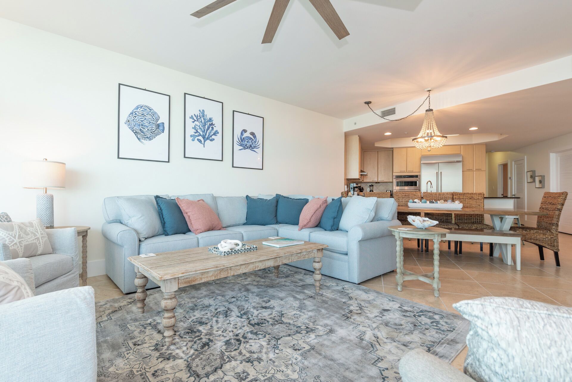 A living room with a light blue sofa, a wooden coffee table, and a patterned rug. There are framed artworks on the wall and a chandelier in the dining area.