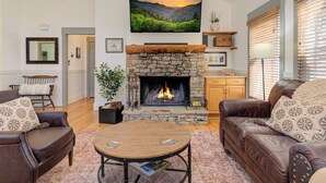Uma sala de estar com lareira de pedra, sofás de couro, uma mesa de centro redonda e um grande quadro de paisagem sobre a lareira.