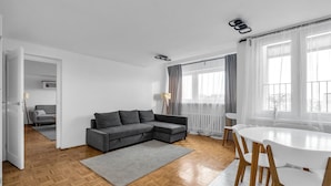 A modern living room with a grey sectional sofa, a white dining table with wooden chairs, and a large window with sheer curtains. - Mokotów Serene Apartment (Warszawa)