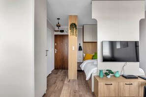 A modern hotel room with a wooden wardrobe, a bed with colorful pillows, a TV mounted on the wall, and a small desk with a vase and a plant. - National Stadium Homely Studio (Warszawa)