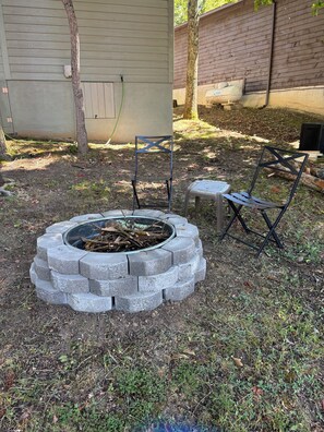 A circular stone fire pit with two metal chairs and a small table, surrounded by grass and trees. - Whispering Pines - a Charming 2-bedroom cabin with WiFi, AC in serene Pipestem (Pipestem)