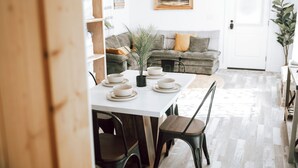 A dining area with a white table set for six, surrounded by dark chairs. A grey sofa with a yellow pillow is against the wall, and a potted plant is on the floor. - The Chicken Coop (Nephi)