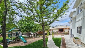 A playground with a slide and climbing structures, a paved pathway, and a building with a garage. - The Chicken Coop (Nephi)