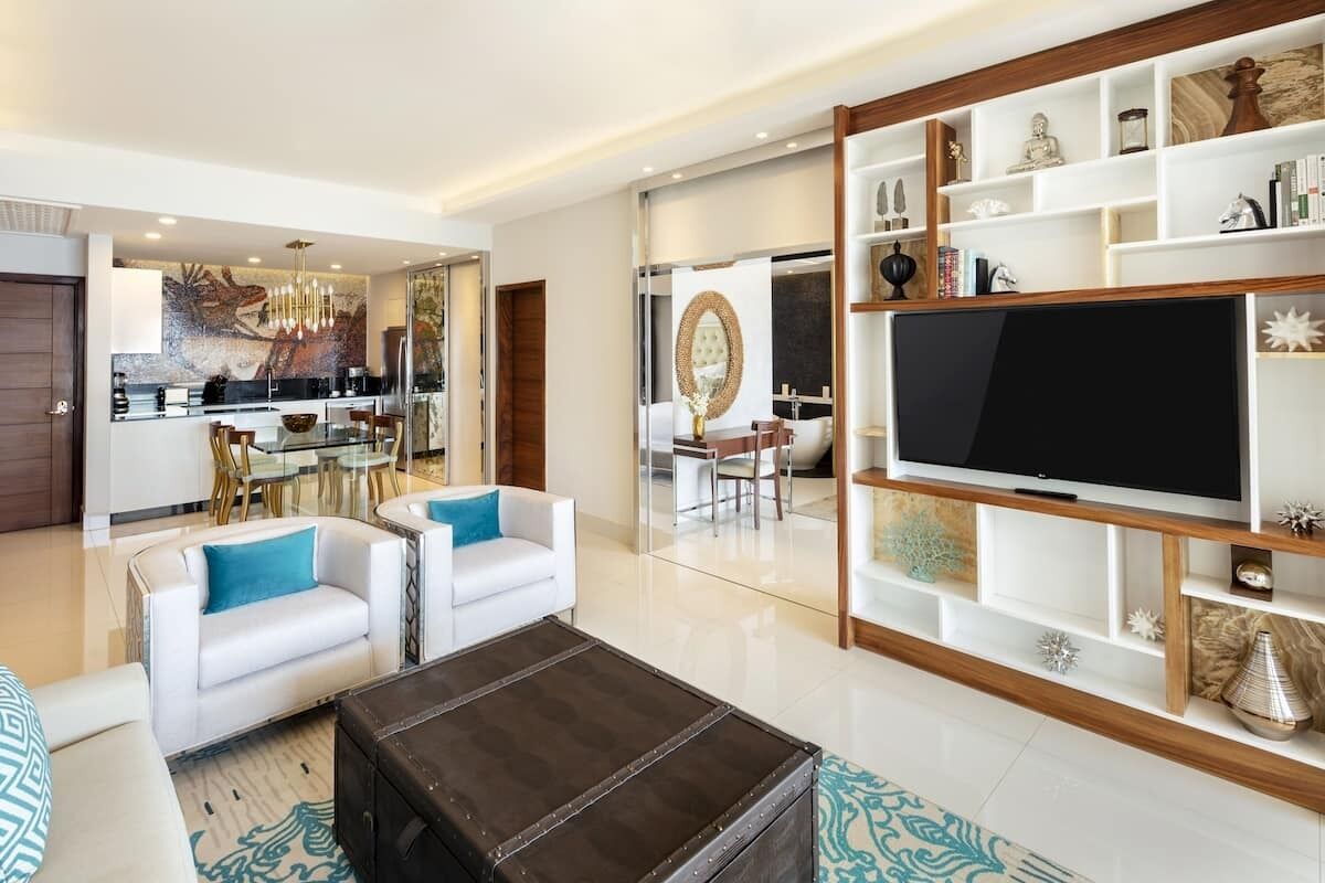 Ein modernes Wohnzimmer mit einem großen Flachbildfernseher, weißen Sofas mit blauen Kissen, einem dunkelbraunen Ottomanen und einem Holz-TV-Schrank mit Dekorationselementen.