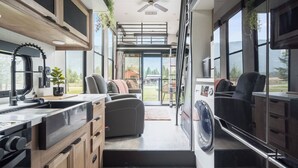 Un intérieur de camping-car compact comprenant un coin cuisine, un espace salon et une grande fenêtre offrant une vue sur l’extérieur.