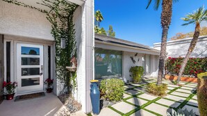 Una casa moderna con fachada blanca, puerta principal con panel de vidrio, pequeño jardín con palmeras y patio con baldosas.