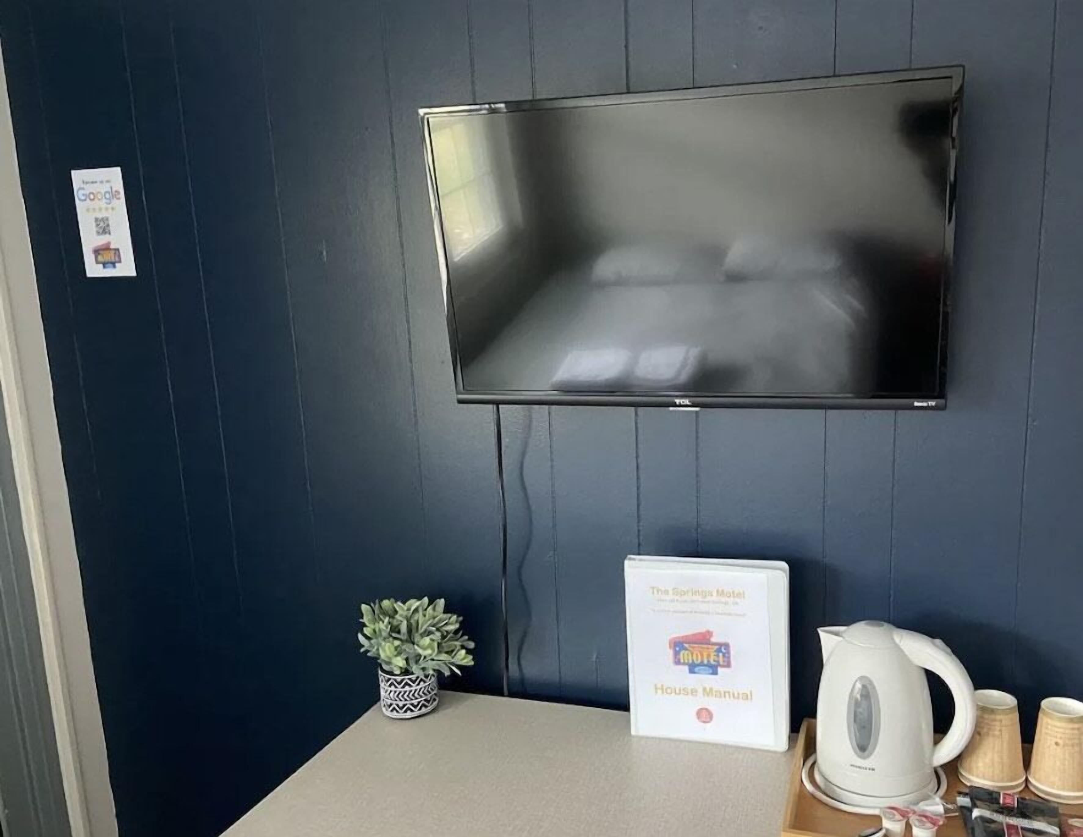 A hotel room with a flat-screen TV mounted on the wall, a small potted plant, a sign with the hotel's name, a kettle, and some cups on the table.