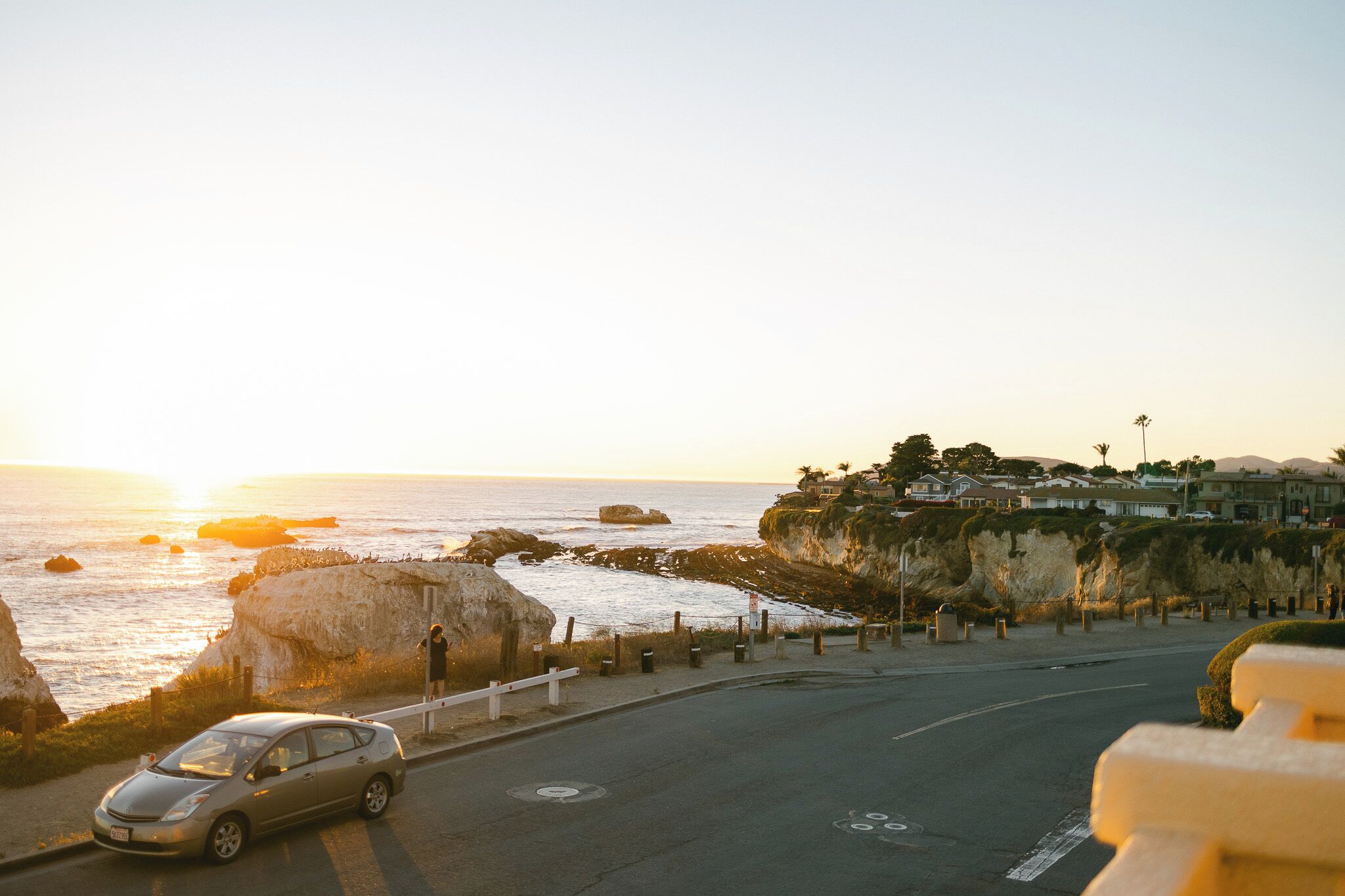 A coastal road with a car, cliffs, and a building complex in the background.