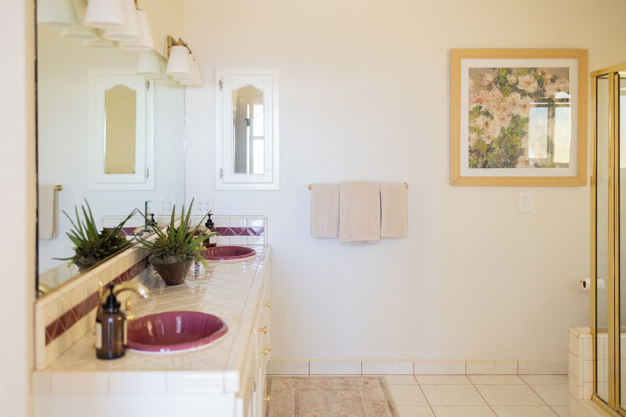 Une salle de bain avec un double lavabo, deux miroirs, une plante en pot et une œuvre d’art florale encadrée accrochée au mur.