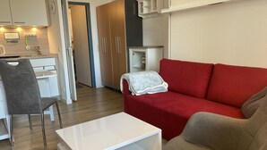 Ein modernes Wohnzimmer mit einem roten Sofa, einem grauen Sessel und einem weißen Couchtisch. Im Hintergrund befindet sich eine Küchenzeile mit Spüle und Herd.