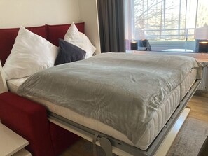 Ein ordentlich bezogenes Bett mit grauer Decke und rotem Kopfteil, ein Nachttisch und ein Fenster mit Vorhängen.
