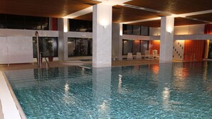 Ein Hallenbad mit klarem Wasser, umgeben von weißen Wänden und Säulen, und einem Sitzbereich mit Stühlen.