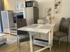 Ein kompakter Wohnbereich mit Küchenzeile, einem Esstisch mit zwei Stühlen, einem Fernseher, einem Kühlschrank und einer Kaffeemaschine.