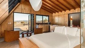 A modern bedroom with a large bed, a wooden ceiling, a dining area, and a view of the desert landscape.