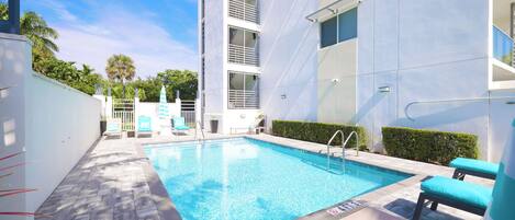 A swimming pool with lounge chairs, a building with balconies, and a clear sky.