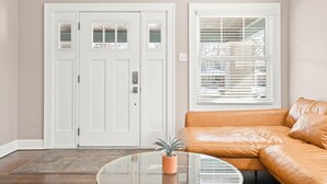 A living room with a beige wall, a white door with glass panels, a window with blinds, and a brown leather sofa. - Adorable Broad Ripple Duplex (Indianapolis)