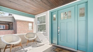 A porch with two wicker chairs, a teal door, and a brick wall. - Adorable Broad Ripple Duplex (Indianapolis)