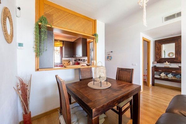 A dining area with a wooden table, chairs, a woven basket, and a kitchen visible through an open doorway.