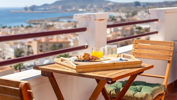 Un balcon avec une table en bois, une chaise, un plateau avec des croissants, un verre de jus d’orange et une tasse.