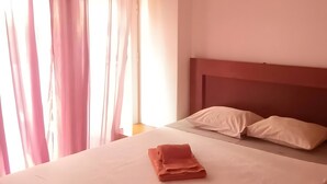 Ein ordentlich bezogenes Bett mit weißer Bettdecke und zwei Kissen, einem Holzkopfende, einer Hängelampe und leichten Vorhängen.