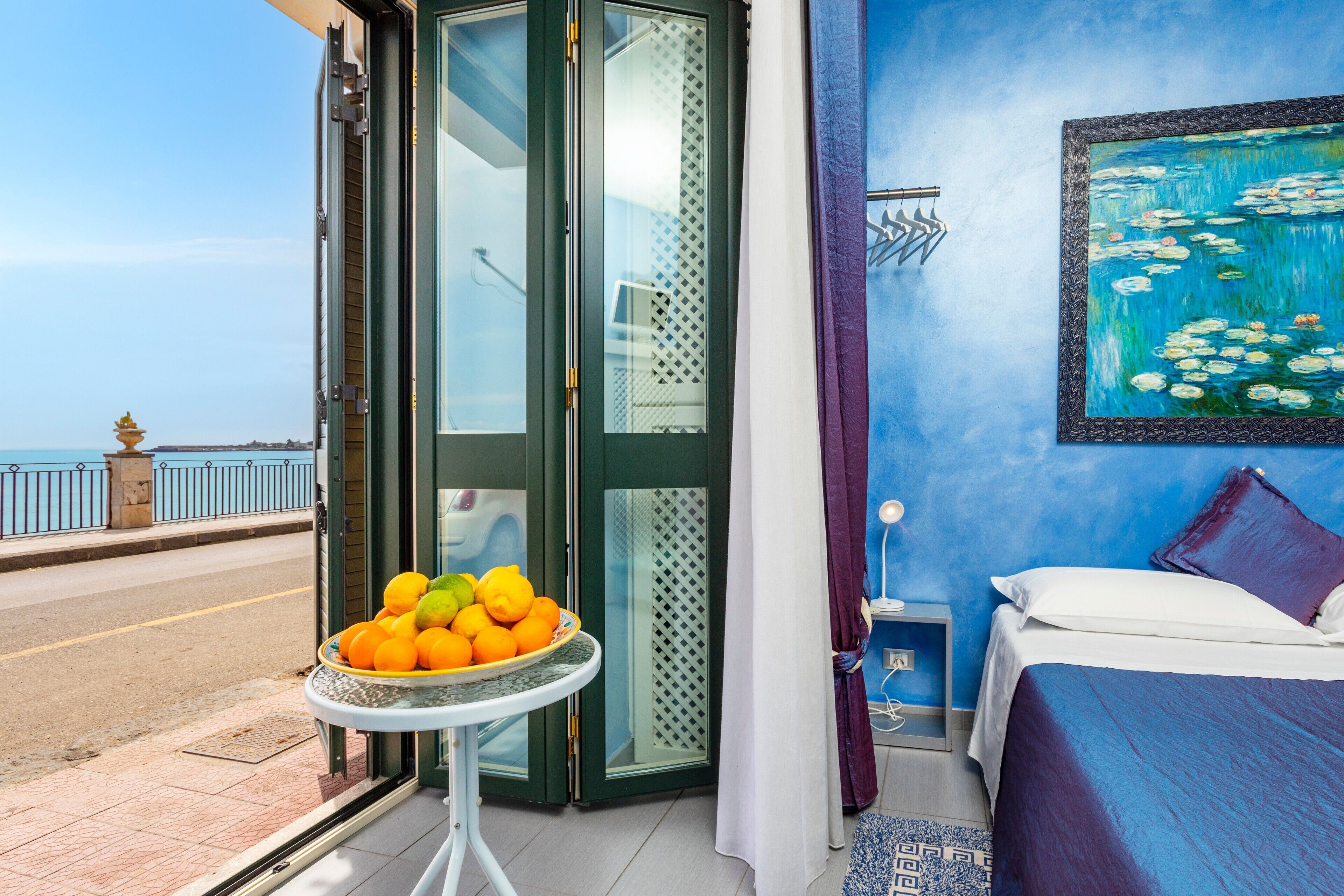 A room with a bed, a painting of water lilies, a fruit bowl on a table, and a view of the sea through a glass door.