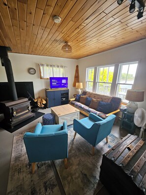 A cozy living room with a wooden ceiling, a fireplace, a sofa, two blue armchairs, a coffee table, and a television.