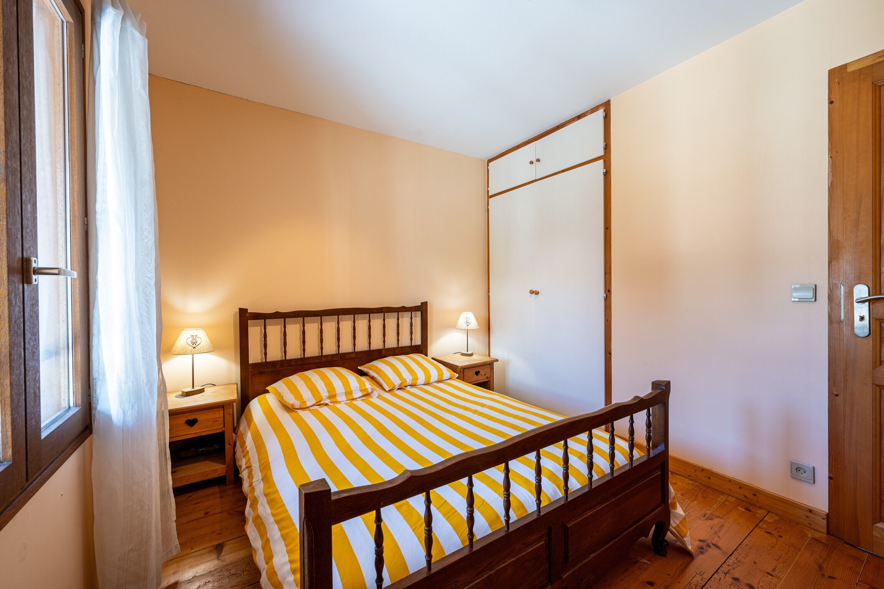 Un dormitorio con una cama de madera, dos mesitas de noche con lámparas, un armario y una ventana con cortinas.