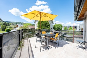 En terrasse med et bord og stole, en parasol og udsigt til huse i baggrunden.