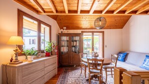 A cozy room with a wooden ceiling, a dining table with chairs, a sofa, a cabinet with books, and a lamp. - Cottage 'Marie Andre' with Private Terrace and Wi-Fi (Entrelacs)