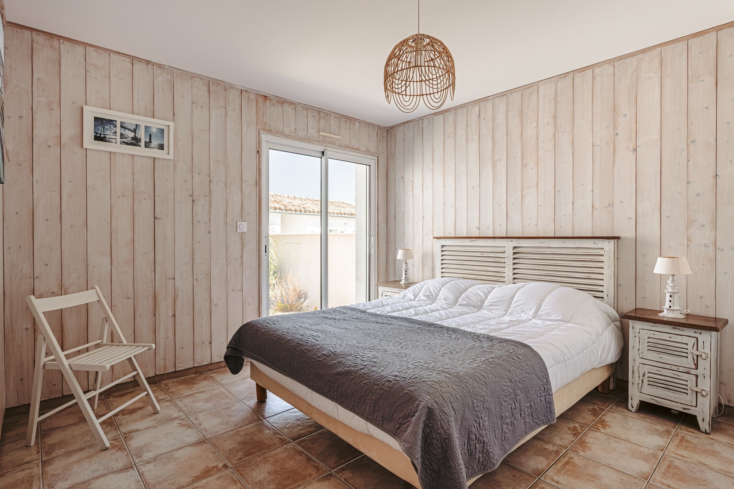 Ein Schlafzimmer mit einem Bett, einem Sessel, einem Nachttisch, einer Lampe, einem Bilderrahmen und einem Fenster.