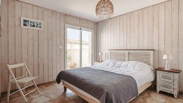 Ein Schlafzimmer mit einem Bett, einem Sessel, einem Nachttisch, einer Lampe, einem Bilderrahmen und einem Fenster.