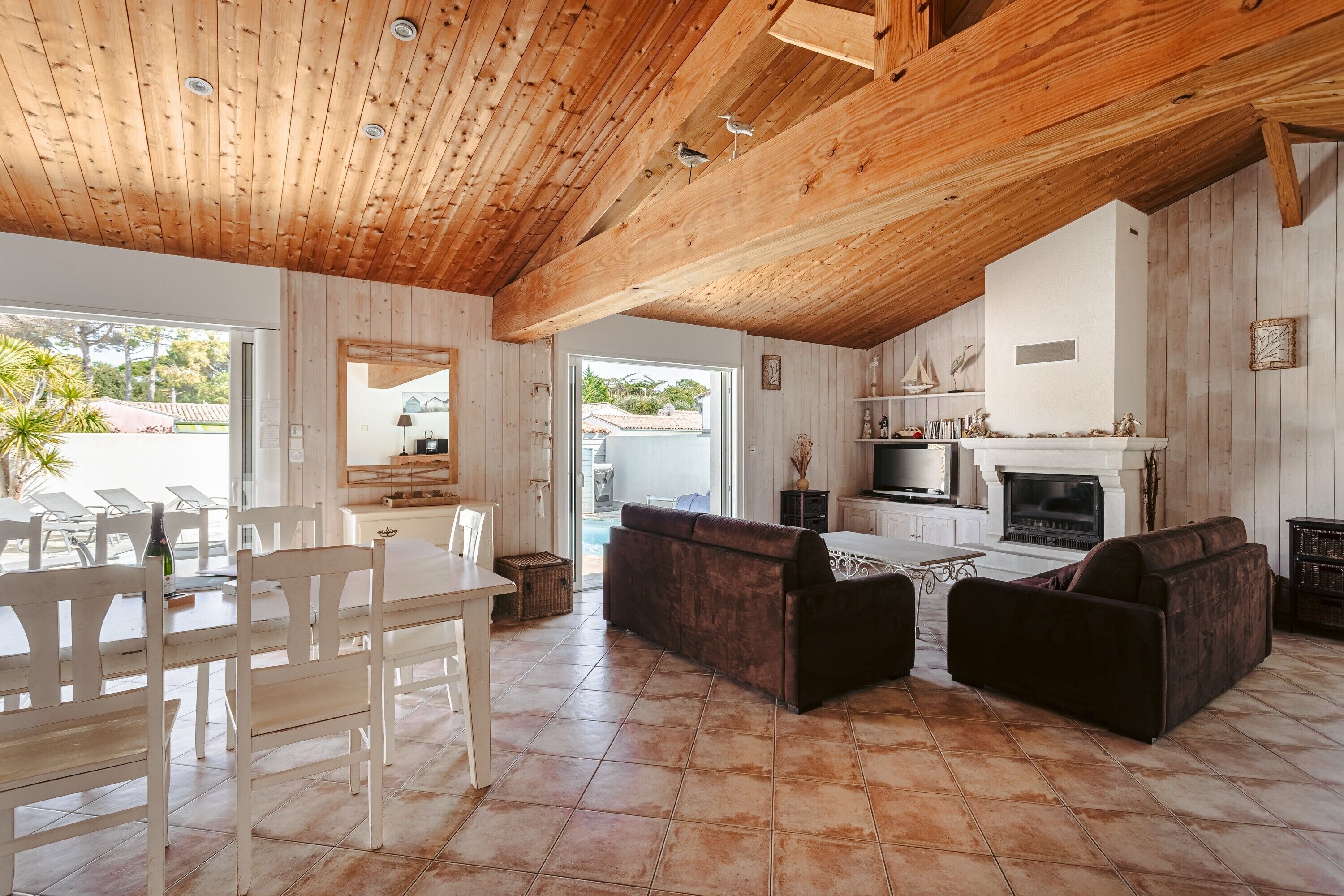Un vaste espace de vie doté d’un plafond en bois, d’une cheminée, d’un téléviseur et d’un coin repas avec des chaises et une table.