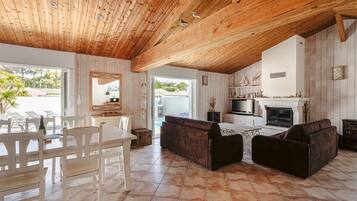 Un vaste espace de vie doté d’un plafond en bois, d’une cheminée, d’un téléviseur et d’un coin repas avec des chaises et une table.