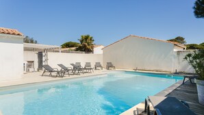 Une piscine avec des chaises longues, une terrasse en bois et un bâtiment blanc doté d’un toit recouvert de tuiles.
