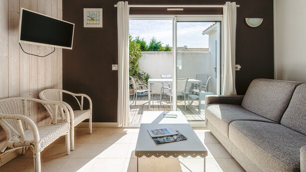 Un salon comprenant un canapé, un fauteuil en osier, un téléviseur à écran plat, une table avec un magazine et une porte-fenêtre coulissante donnant sur un espace extérieur avec une table et des chaises.