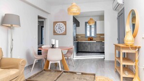 En moderne stue med sofa, spisebord med stole, sofabord i træ, en hængende vævet lampe, vægur og et køkken, der kan ses igennem en åben dør.
