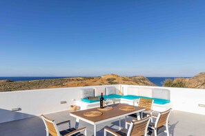 Una terraza exterior con una mesa, sillas y un sofá con cojines, con vistas a un paisaje costero.