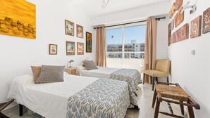 A hotel room with two beds, a chair, a small table, artwork on the walls, and a view of buildings through the window. - Apartment 'Del Mar' with Sea View, Wi-Fi and Air Conditioning (Marbella)