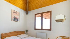 Ein Schlafzimmer mit einem hölzernen Bett, einem Fenster mit Jalousien, einem wandmontierten Spiegel und einem Korbstuhl mit orangefarbenem Kissen.